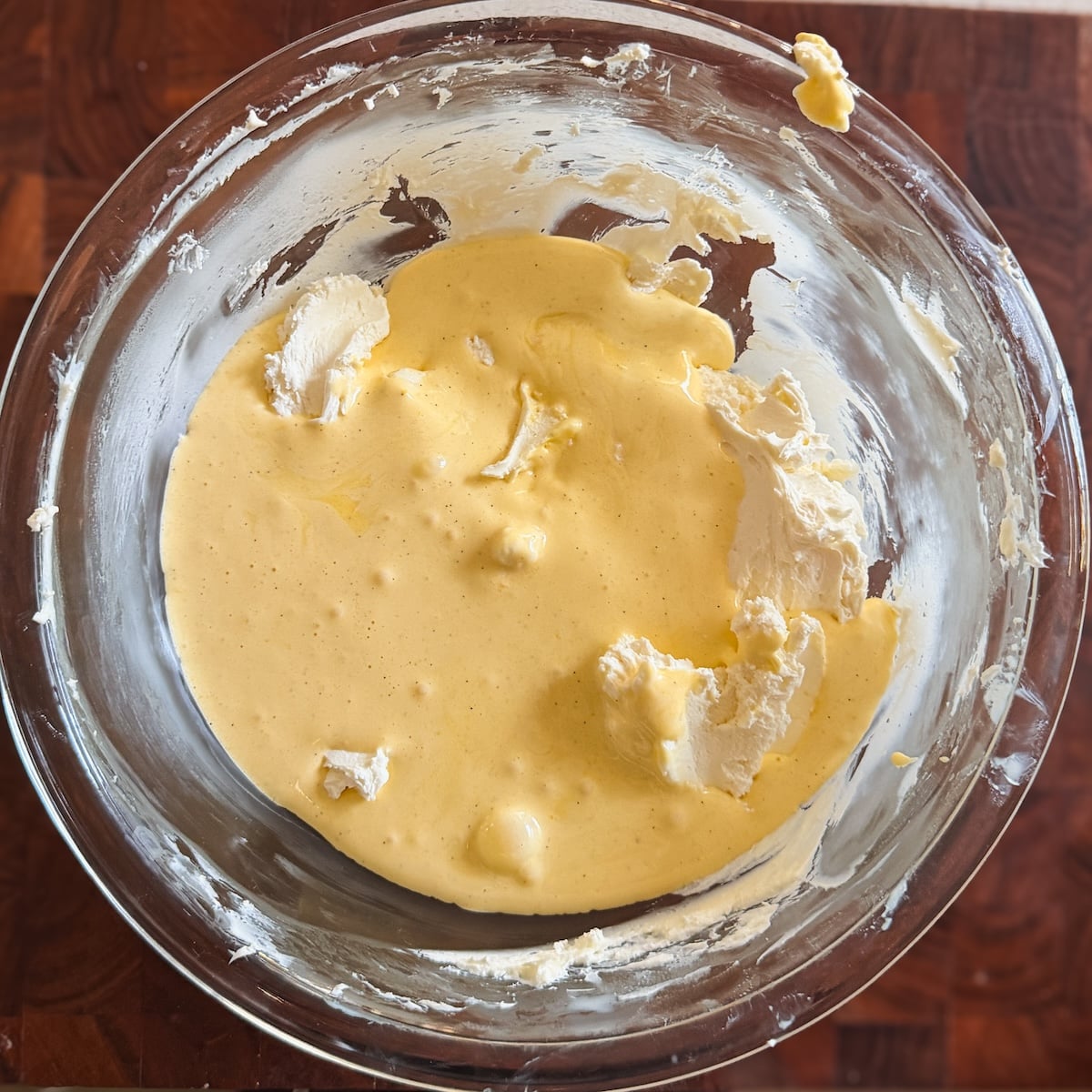 A clear glass bowl containing a creamy, yellow batter with some white lumps, sitting on a wooden surface. Ingredients appear to be partially mixed.
