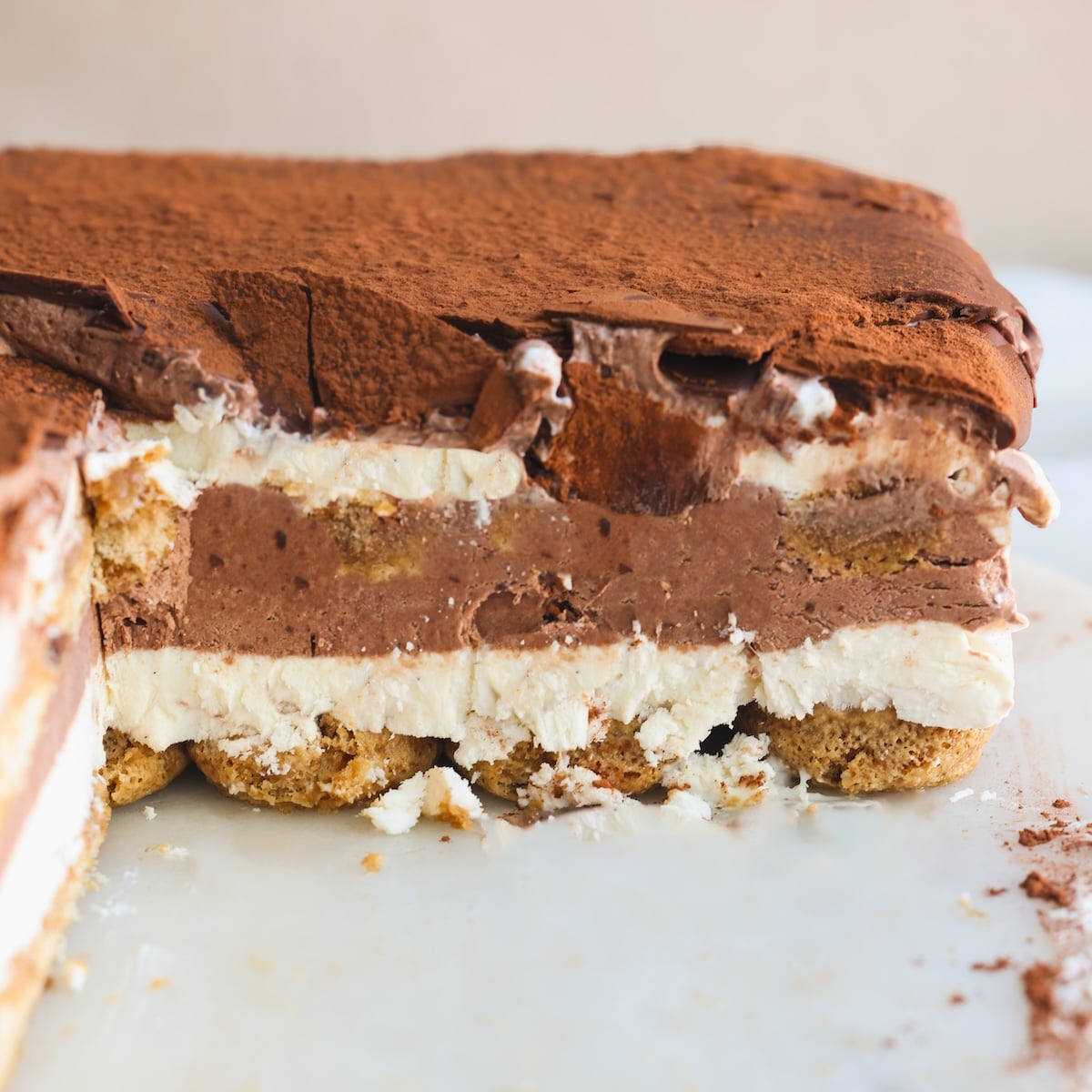 A close-up of a sliced layered dessert with vanilla ice cream, chocolate ice cream, and ladyfingers, dusted with cocoa powder on top, sitting on a white surface.
