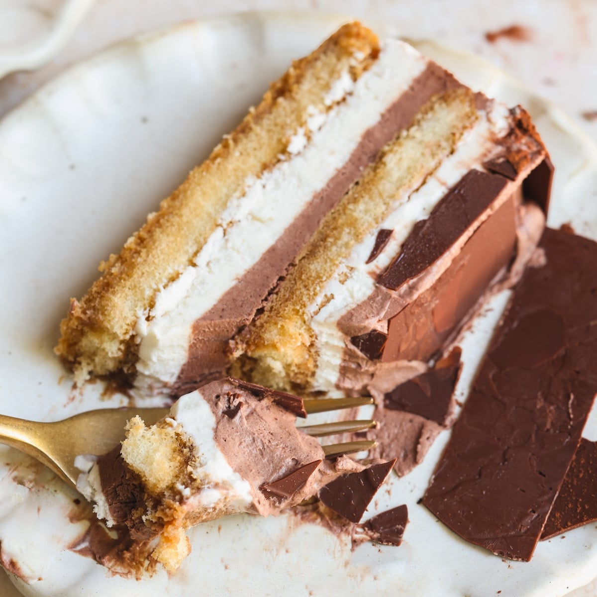 A slice of layered tiramisu ice cream cake on a white plate, with a fork taking a bite. The cake has alternating layers of chocolate and vanilla ice cream, topped with a chocolate coating.