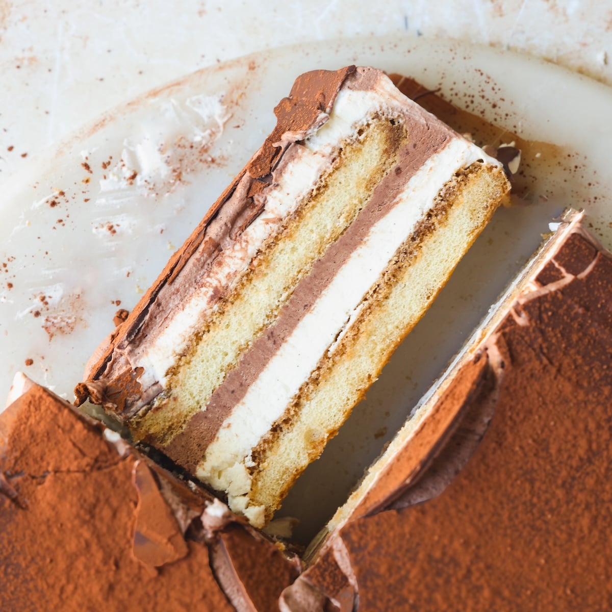 A close-up of a sliced layered tiramisu ice cream cake, showing alternating layers of creamy filling and sponge cake, topped with cocoa powder and chocolate coating on a white surface.