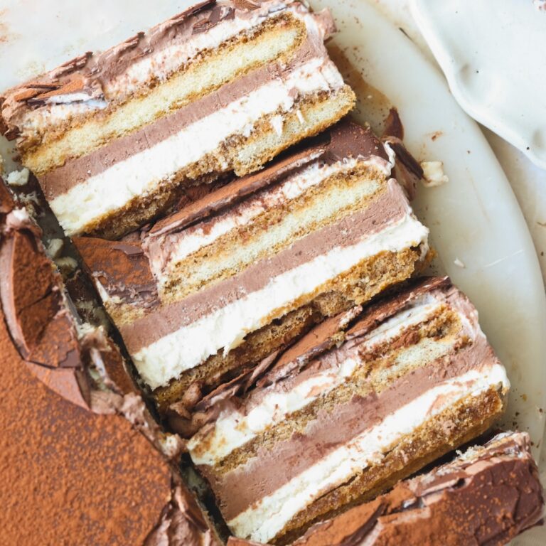 Three slices of layered tiramisu ice cream cake with visible layers of cream, chocolate, and espresso-soaked ladyfingers, placed on a white plate with some cocoa powder on the side.