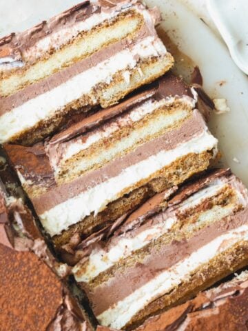 Three slices of layered tiramisu ice cream cake with visible layers of cream, chocolate, and espresso-soaked ladyfingers, placed on a white plate with some cocoa powder on the side.