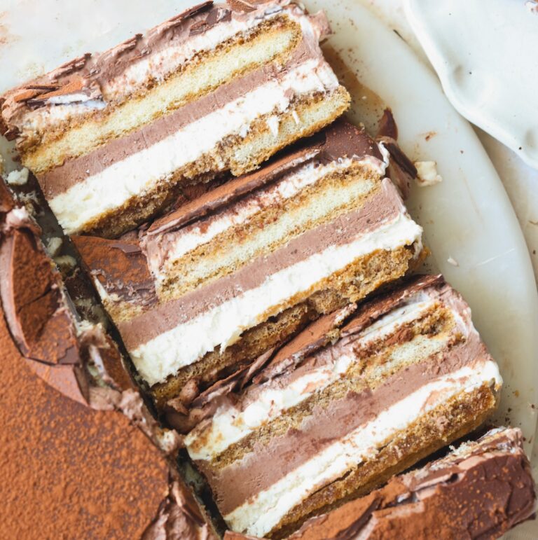 Three slices of layered tiramisu ice cream cake with visible layers of cream, chocolate, and espresso-soaked ladyfingers, placed on a white plate with some cocoa powder on the side.