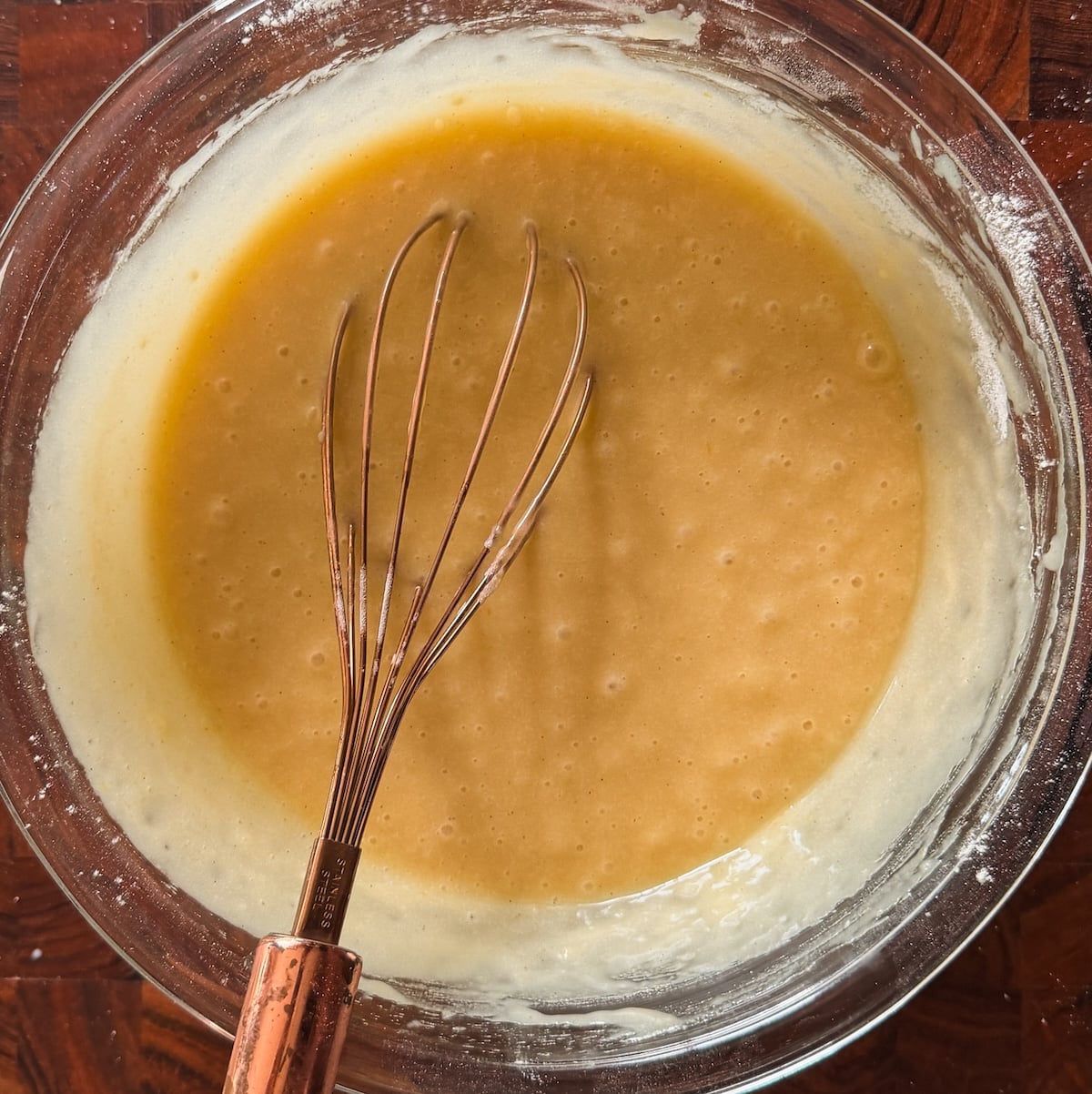 A glass bowl filled with smooth, light brown batter sits on a wooden surface. A metal whisk with a copper handle rests in the mixture, showing signs of recent stirring. Batter is clinging to the sides of the bowl.
