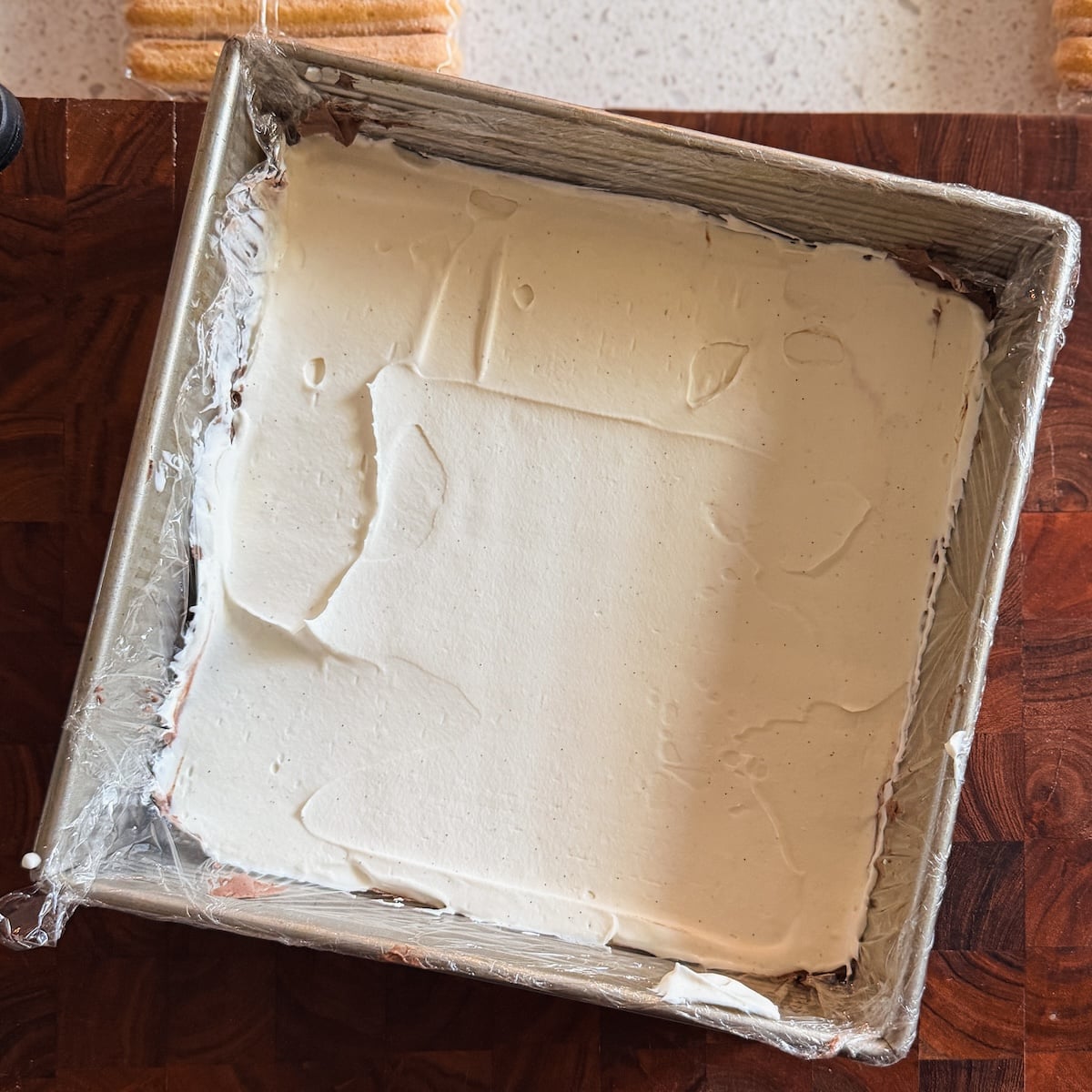 A square metal baking pan lined with plastic wrap, filled with a thick, smooth layer of white ice cream, on a wooden surface.