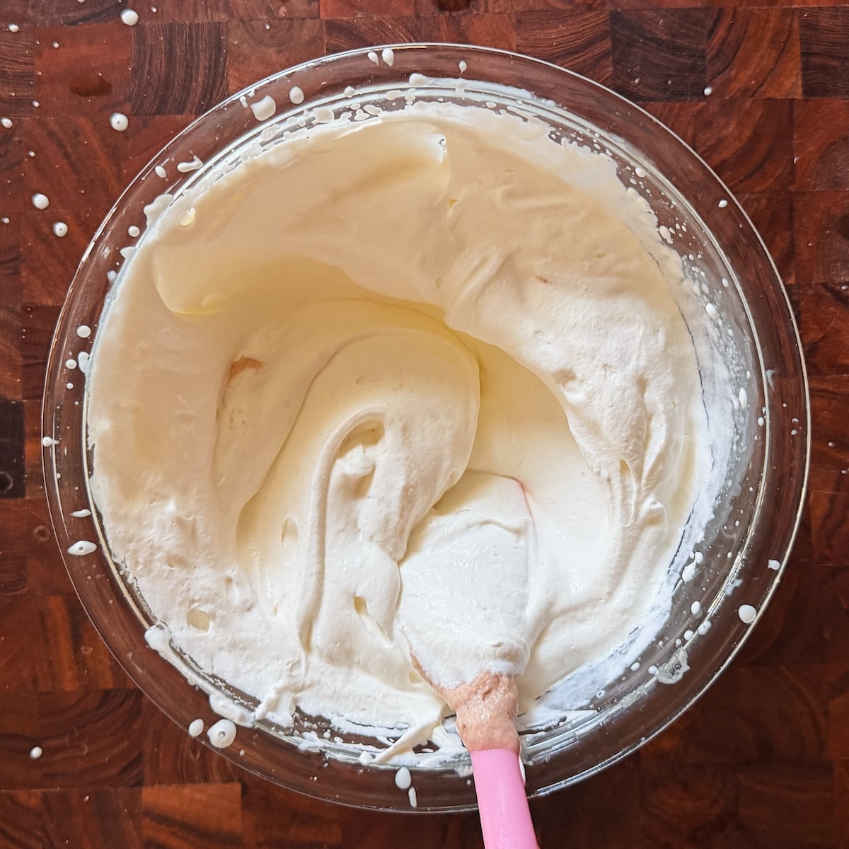 A glass bowl filled with whipped ice cream being mixed with a pink spatula, resting on a wooden surface with some cream splatters around.