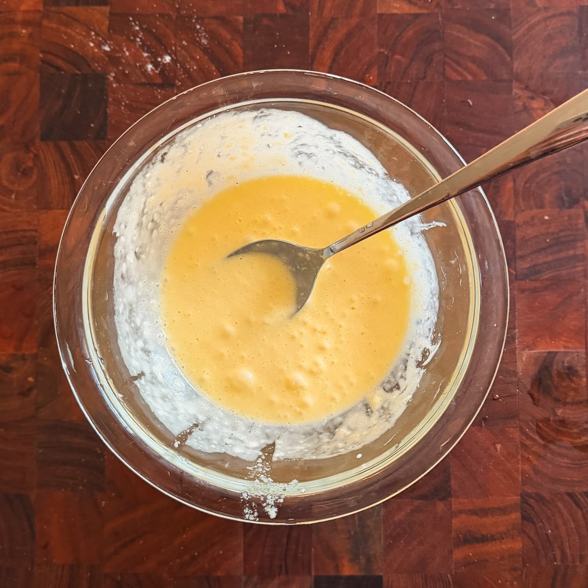 A glass bowl sits on a wooden surface, filled with a pale yellow, slightly bubbly mixture. A metal spoon rests in the mixture, indicating it has just been stirred. Some batter clings to the sides of the bowl.