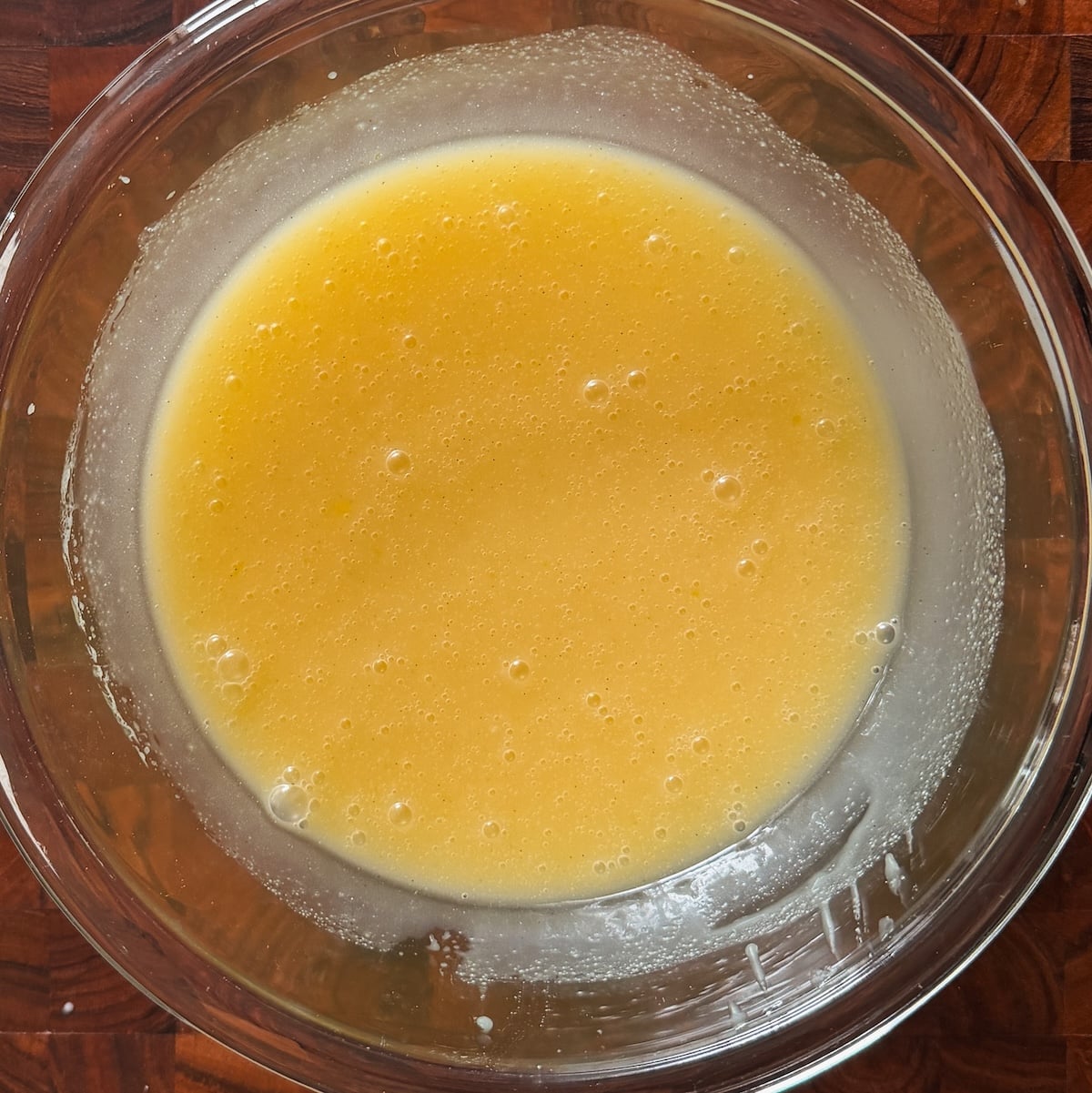 A glass bowl filled with a smooth, yellow liquid mixture sits on a wooden surface. Small bubbles are visible on the surface of the mixture.