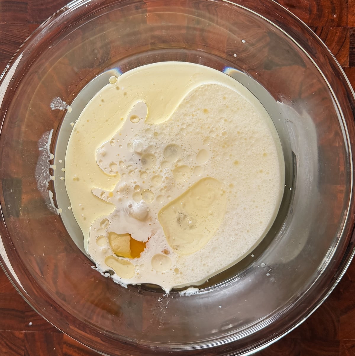 A clear glass bowl containing wet ingredients and sugar on a dark wooden surface, with the ingredients not yet mixed together.