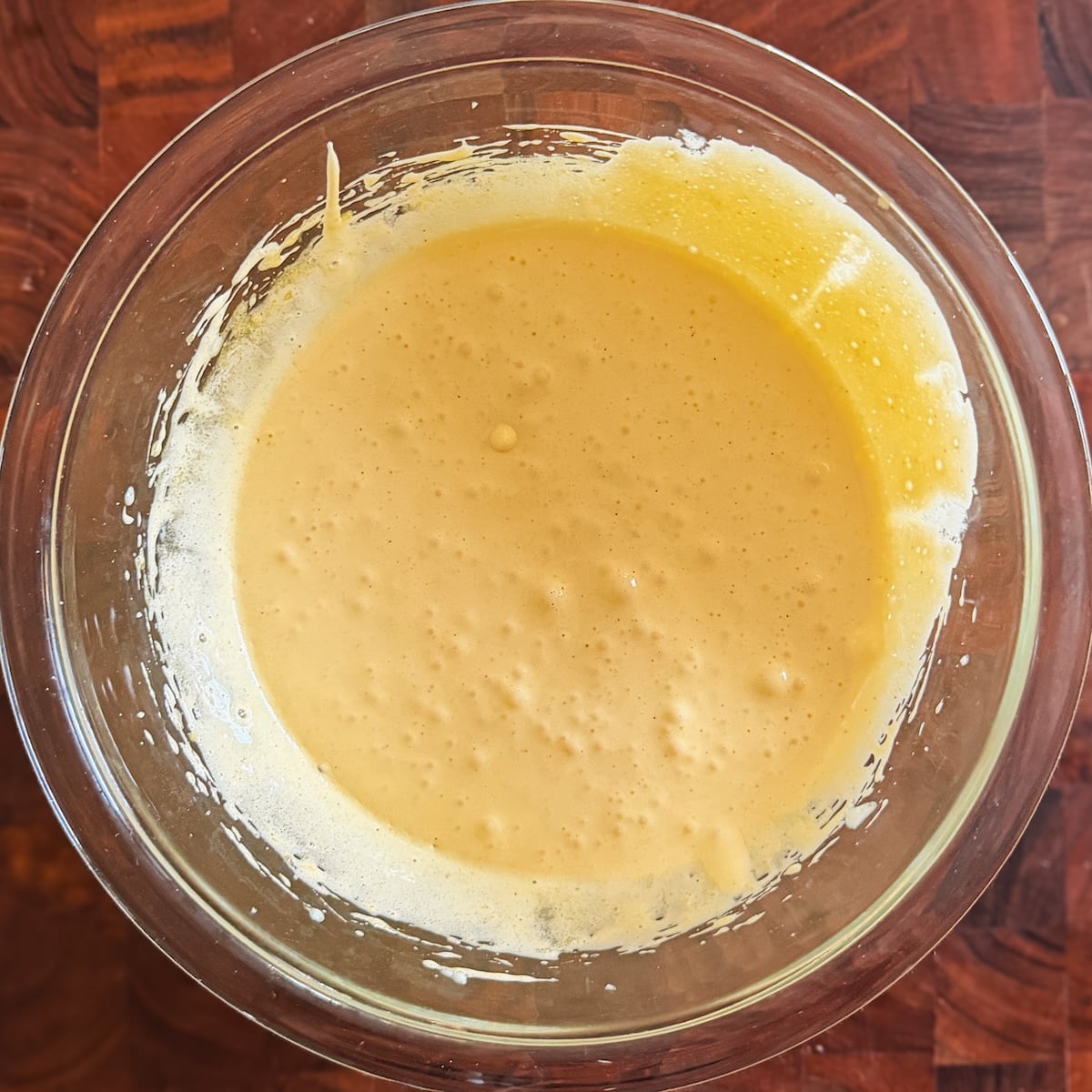 A glass bowl filled with smooth, pale yellow mixture sits on a wooden surface. The batter has a slightly airy texture with small bubbles visible on the surface.