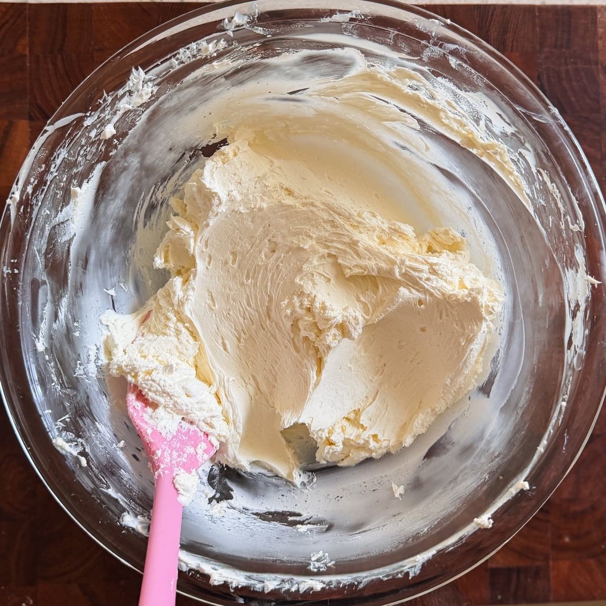 A glass mixing bowl containing creamy, whipped mascarpone, with a pink spatula resting in the mixture on a wooden surface.