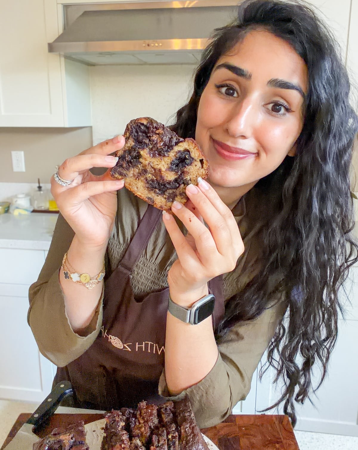 A woman with long dark hair smiles at the camera while holding up a thick slice of brownie banana bread in a kitchen. She wears a brown apron, a smartwatch, and gold bracelets. The rest of the loaf sits on a cutting board.