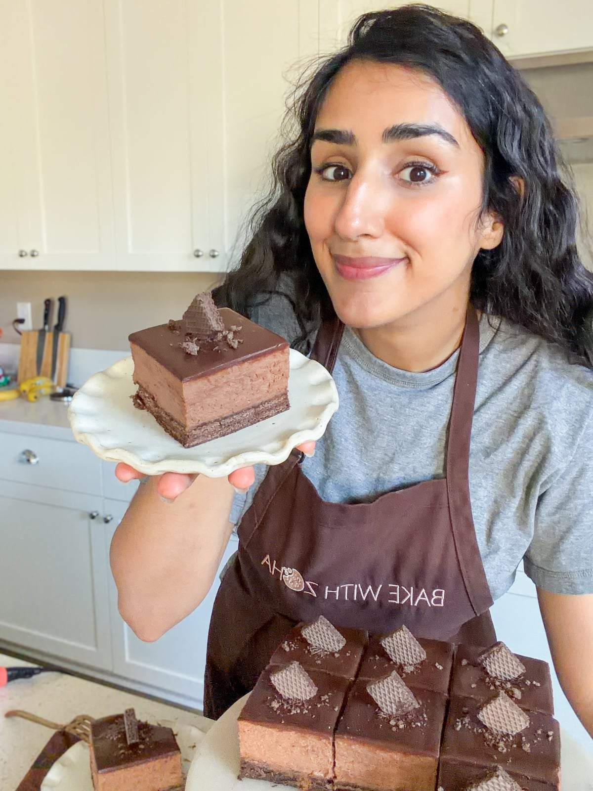 A woman wearing a brown apron and gray shirt smiles while holding a plate with a slice of layered chocolate mousse cake. More slices of the same cake are on the counter in front of her in a bright kitchen.