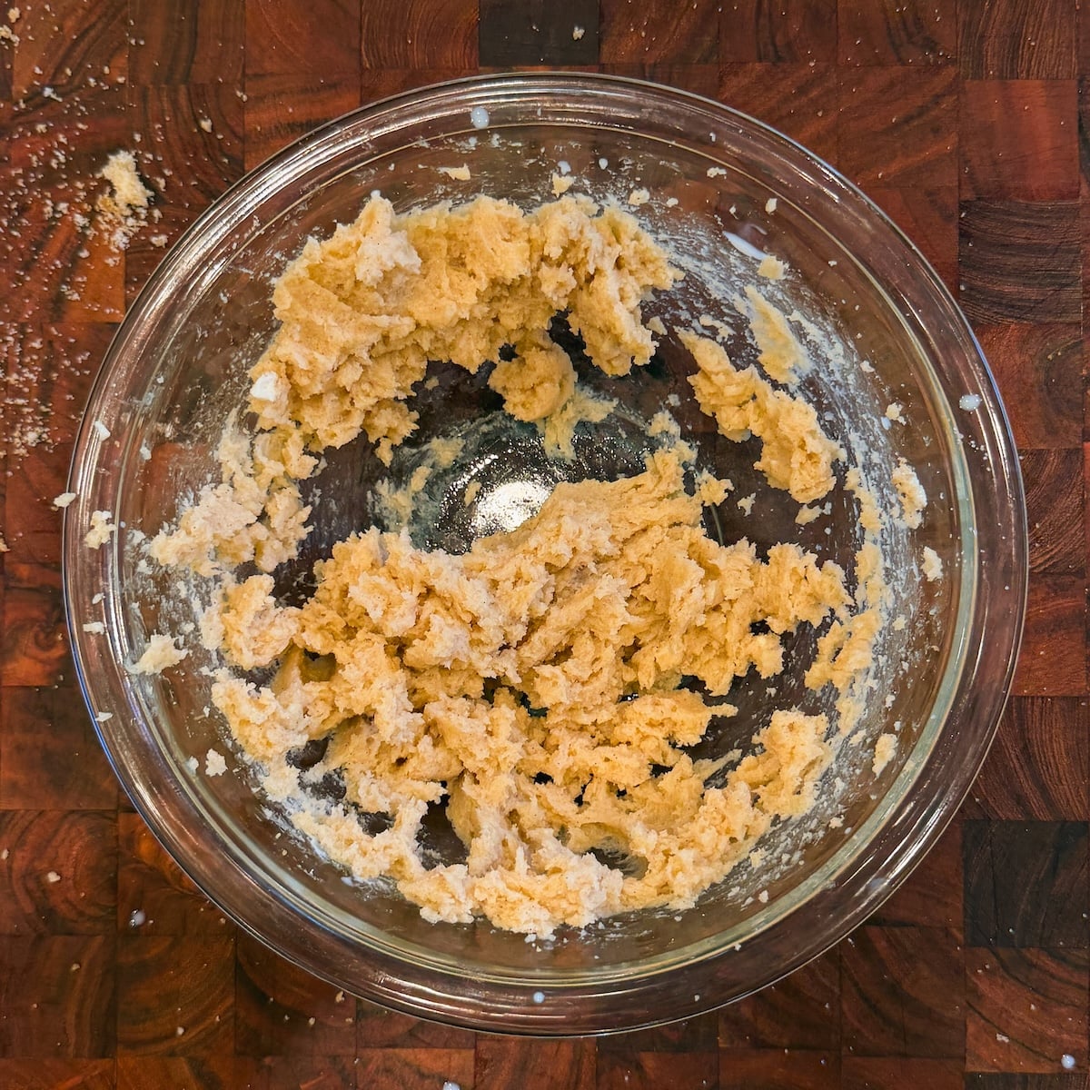 A glass bowl with partially mixed cookie dough sits on a wooden surface. Bits of flour and dough are scattered around the bowl's edge and on the cutting board underneath.