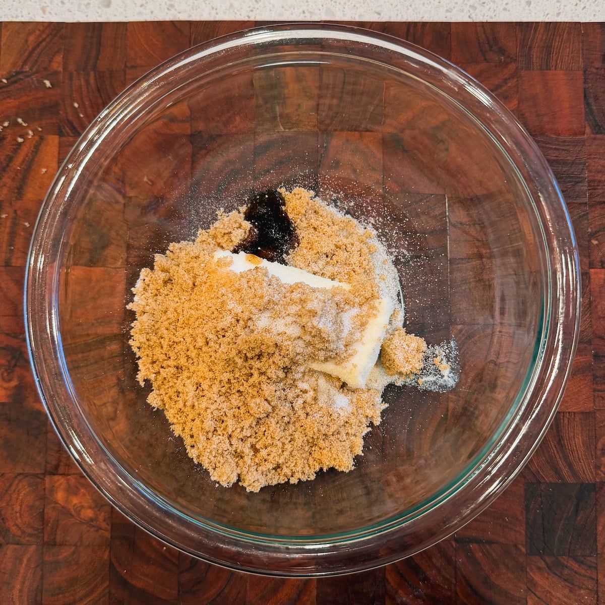A glass bowl on a wooden surface contains brown sugar, white sugar, butter, and a splash of vanilla extract, ready for mixing.