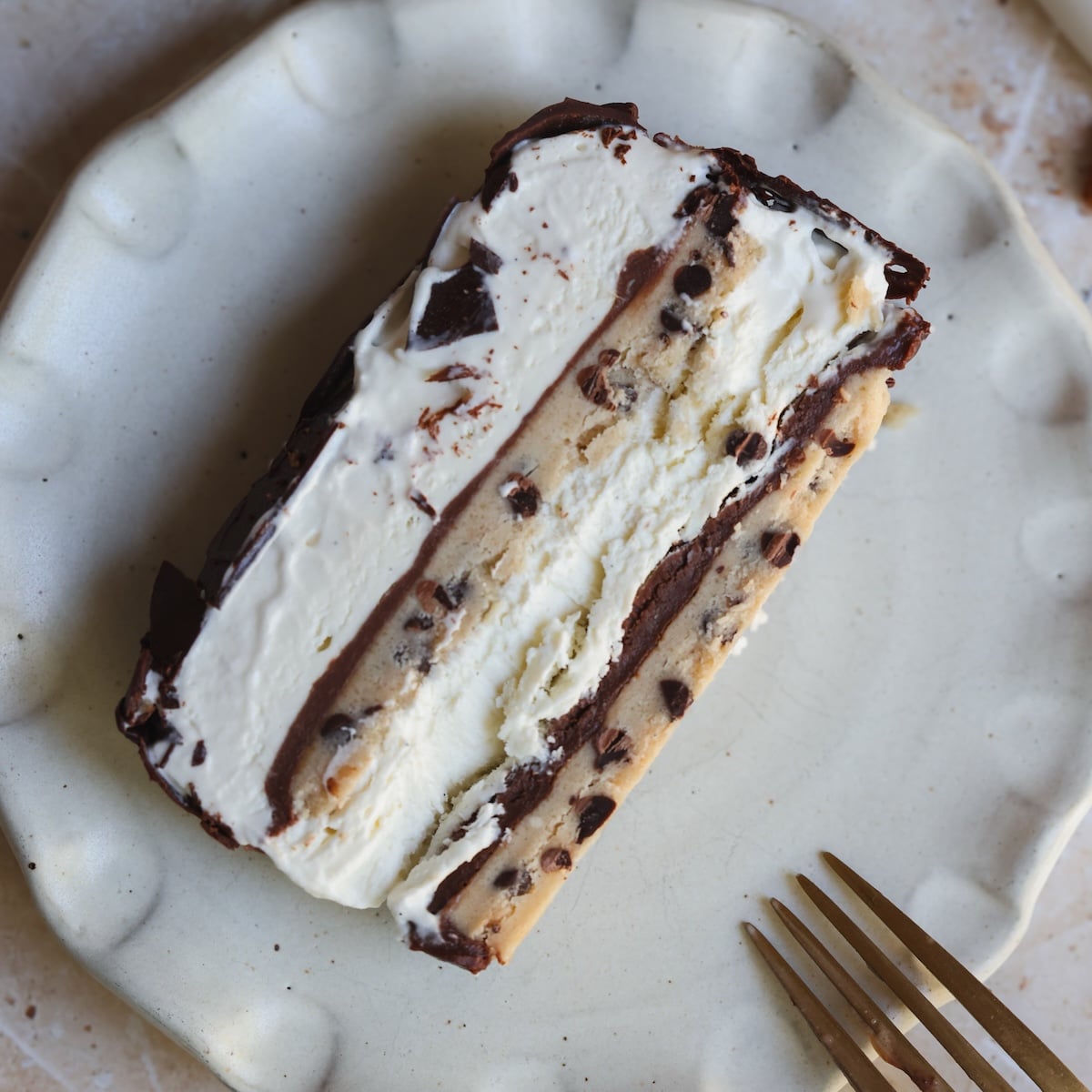 A slice of ice cream cake on a white plate, with layers of vanilla ice cream, chocolate, and chocolate chip cookie dough. A gold fork sits nearby on the plate.