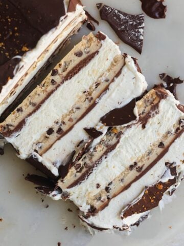Three slices of layered ice cream cake with chocolate and cookie dough layers, topped with a chocolate coating, are on a white surface with scattered chocolate pieces.
