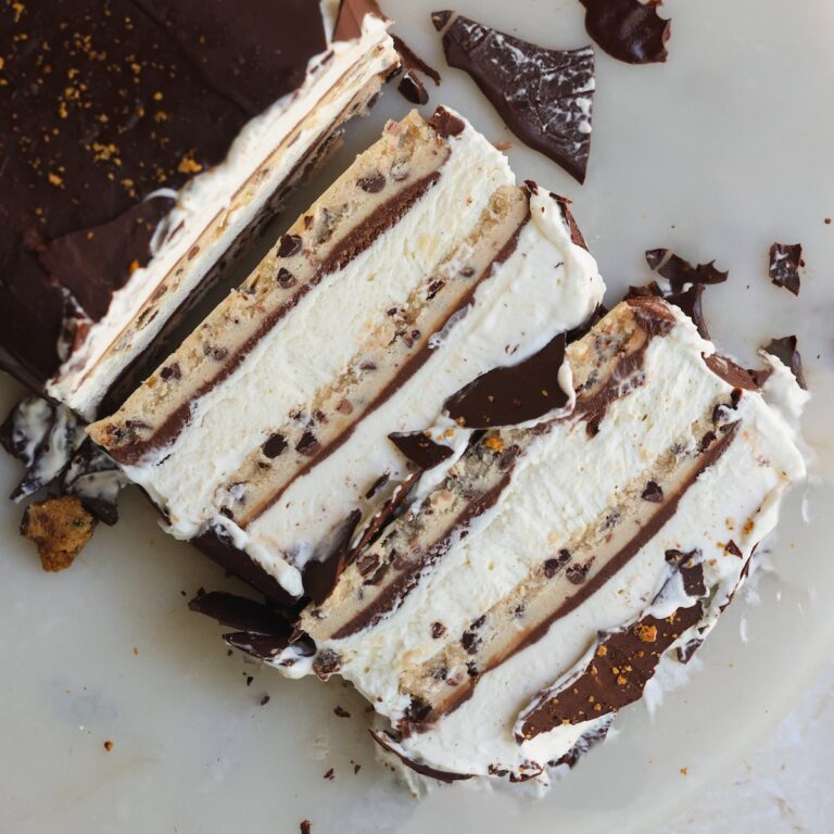 Three slices of layered ice cream cake with chocolate and cookie dough layers, topped with a chocolate coating, are on a white surface with scattered chocolate pieces.
