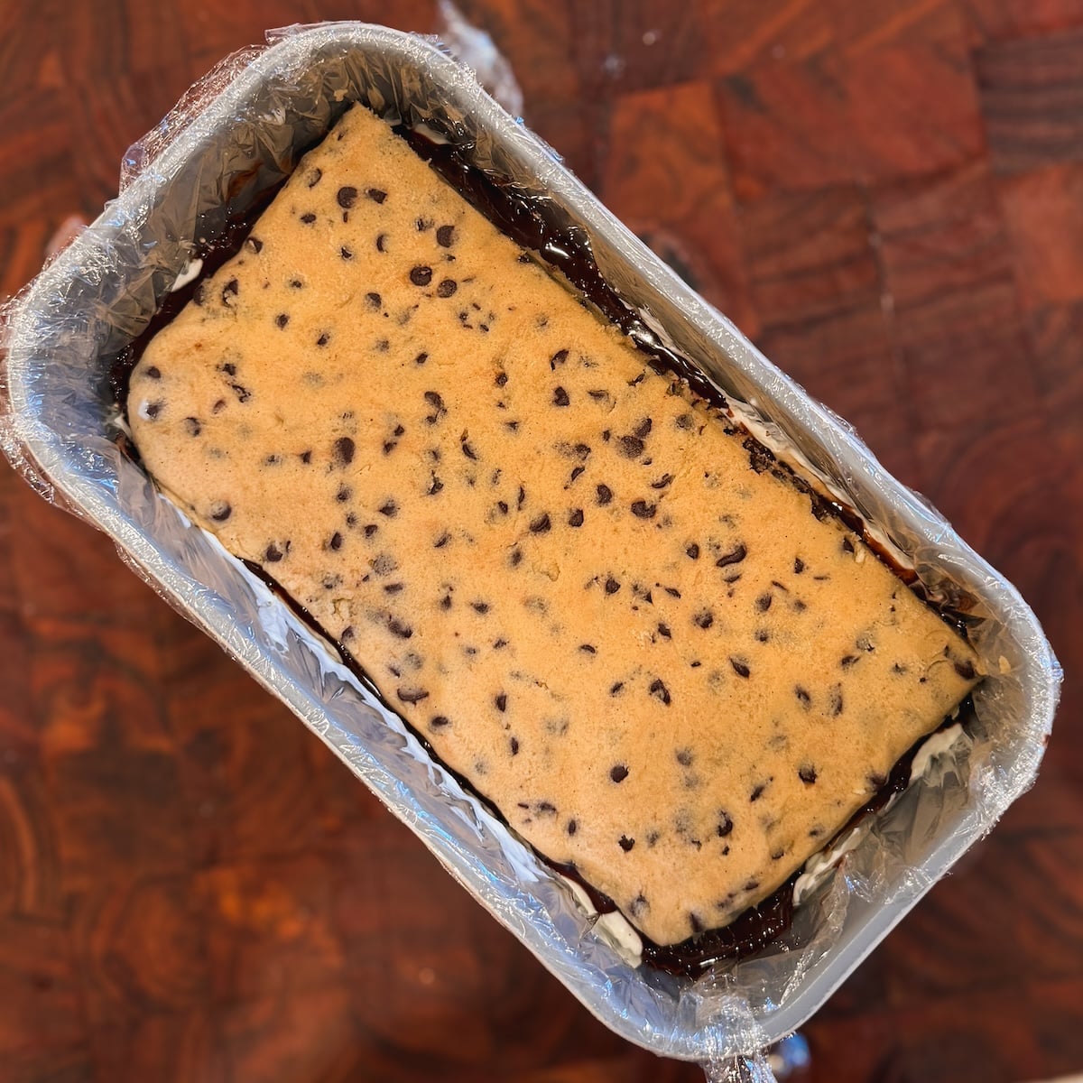 A rectangular loaf pan lined with plastic wrap holds a layer of cookie dough with mini chocolate chips, sitting on a wooden surface.