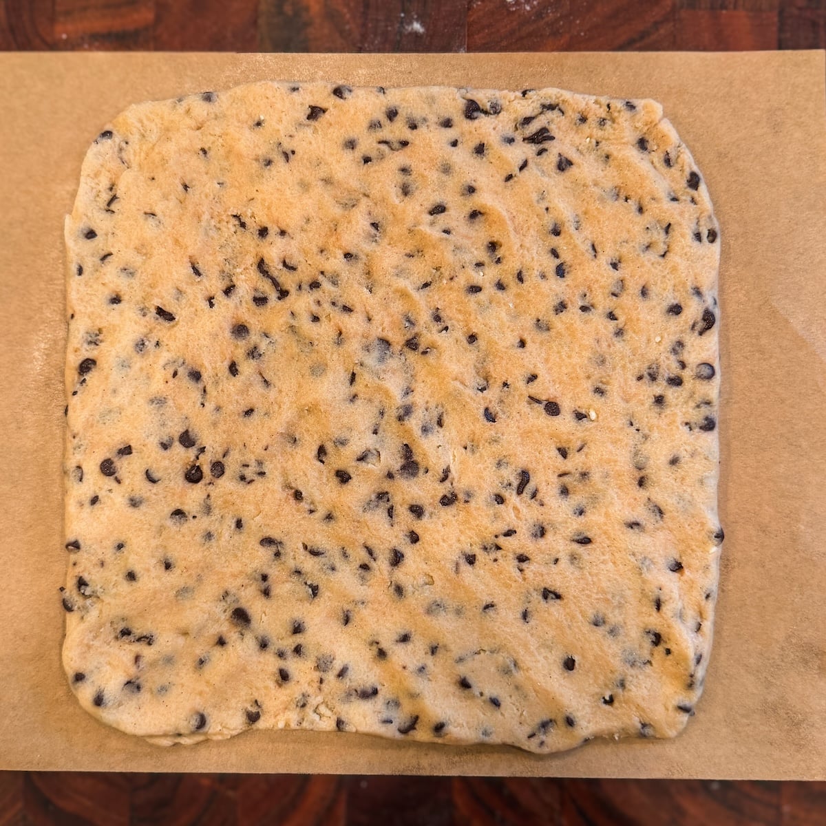Square sheet of chocolate chip cookie dough rolled out on brown parchment paper, resting on a wooden surface.