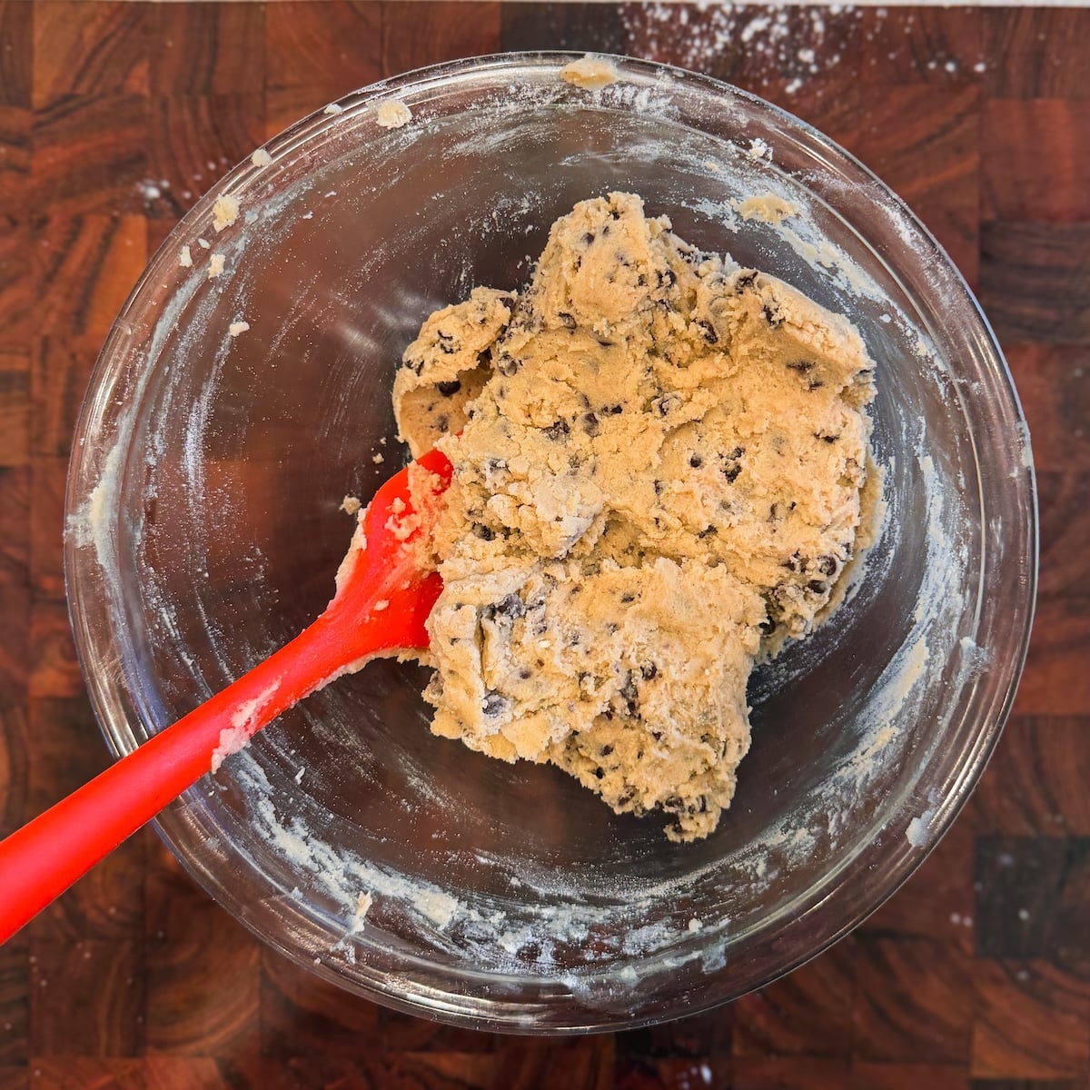 A glass bowl with chocolate chip cookie dough and a red spatula sits on a wooden surface, surrounded by a dusting of flour.