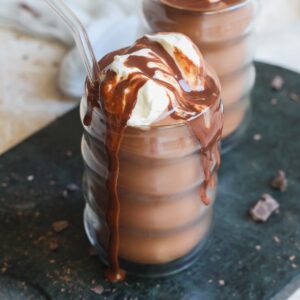 A glass of frozen hot chocolate topped with whipped cream and chocolate sauce, with a clear straw, sits on a dark surface alongside chocolate pieces.