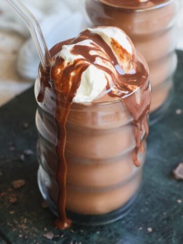 A glass of frozen hot chocolate topped with whipped cream and chocolate sauce, with a clear straw, sits on a dark surface alongside chocolate pieces.