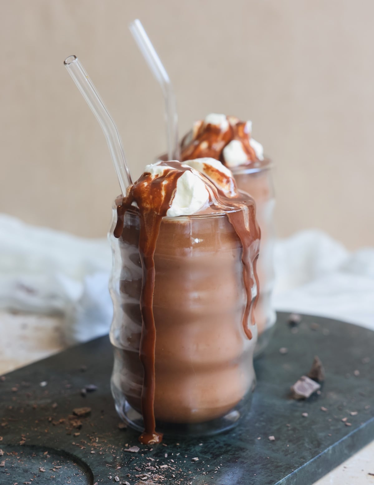 Two iced chocolate drinks in wavy glasses topped with whipped cream and chocolate sauce, with clear straws. Drinks are set on a dark surface with chocolate pieces scattered around.