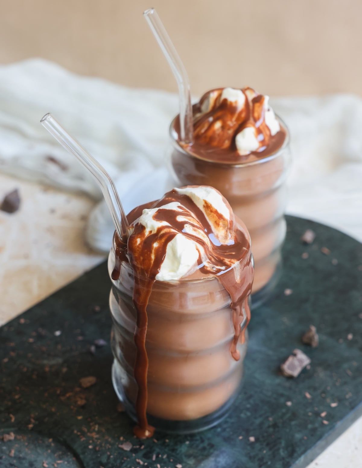Two glasses of frozen hot chocolate topped with whipped cream and chocolate syrup, served with clear straws. The drinks are on a dark tray with some chocolate pieces scattered around.