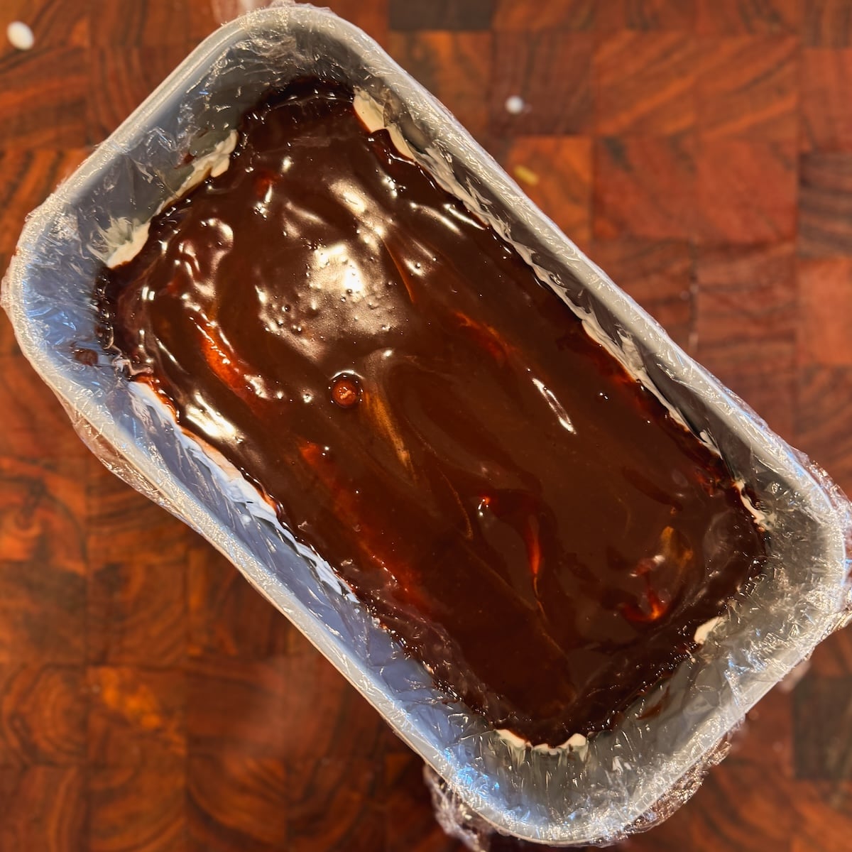 A rectangular pan lined with plastic wrap contains a dessert topped with a glossy chocolate layer, set on a wooden surface.