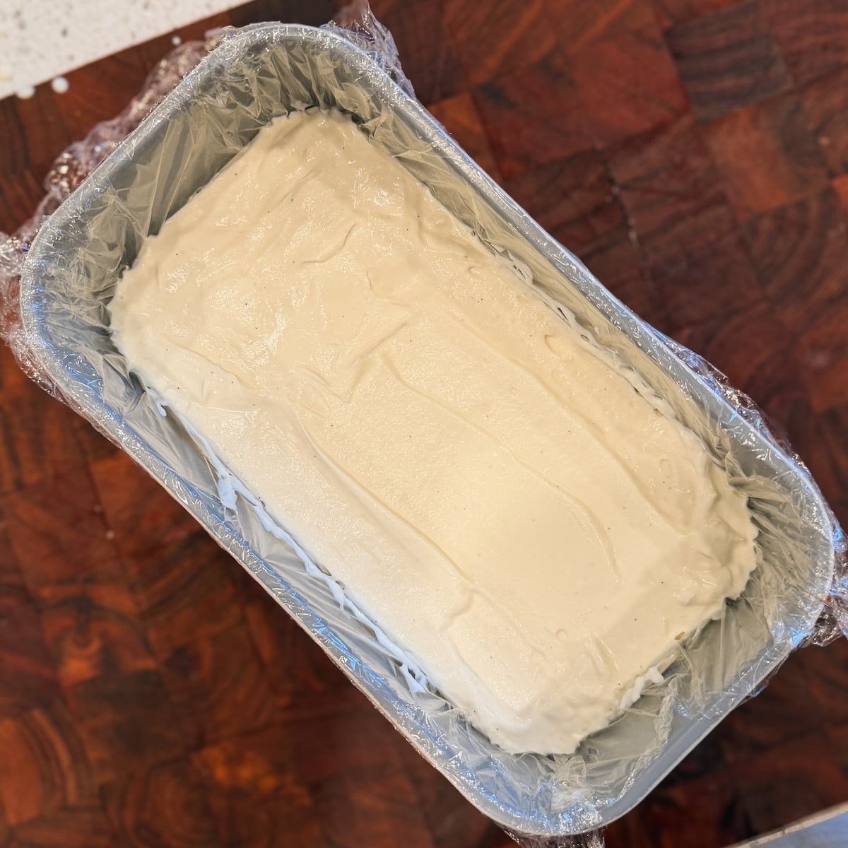 A rectangular pan lined with plastic wrap is filled with a smooth, creamy white ice cream mixture, sitting on a dark wooden surface.