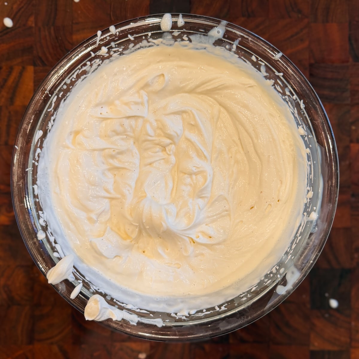 A glass bowl filled with creamy, whipped mixture sits on a wooden surface, with some of the mixture splattered on the bowls rim and a few drops on the table.