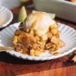 A slice of apple cinnamon bread pudding topped with vanilla ice cream and toffee sauce sits on a white scalloped plate, with a fork and apple slices in the background.