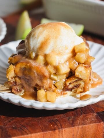 A slice of apple cinnamon bread pudding topped with vanilla ice cream and toffee sauce sits on a white scalloped plate, with a fork and apple slices in the background.