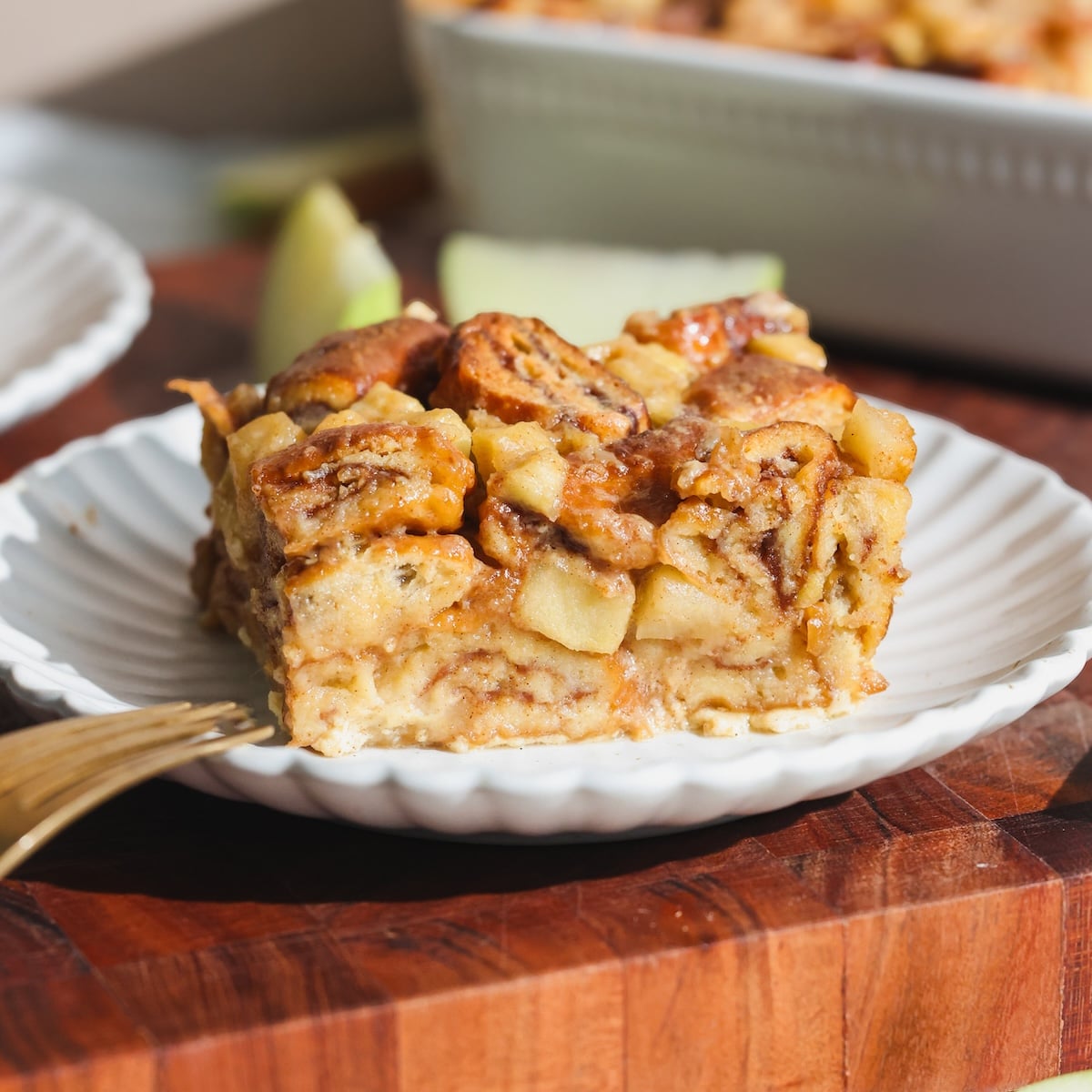 A slice of apple cinnamon bread pudding with visible apple chunks and cinnamon on a white scalloped plate, placed on a wooden surface next to a gold fork.