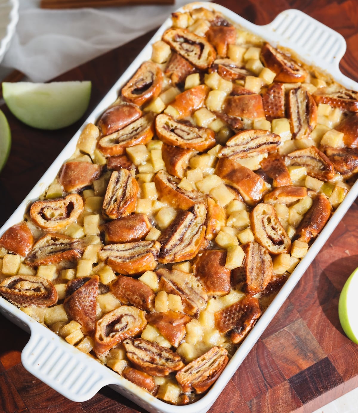 A white baking dish filled with an apple pie cinnamon roll bread pudding. It has diced apples and cinnamon swirl bread, baked to a golden brown. Sits on a wooden surface with green apple slices nearby.