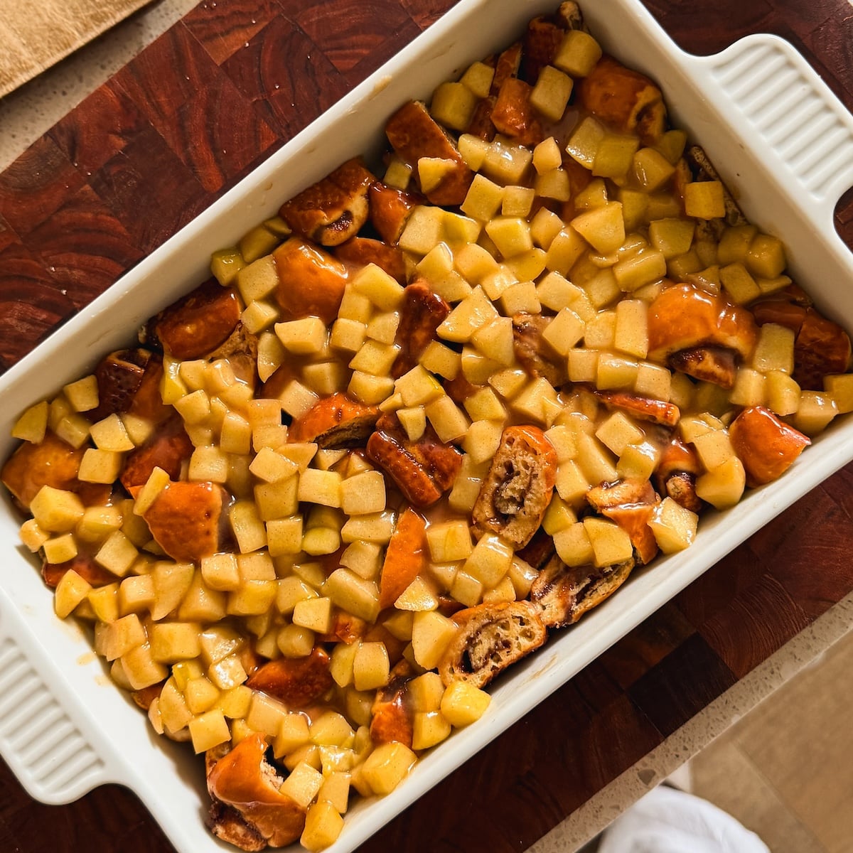 A white baking dish filled with cinnamon bread topped with chopped cooked apples sits on a wooden cutting board.