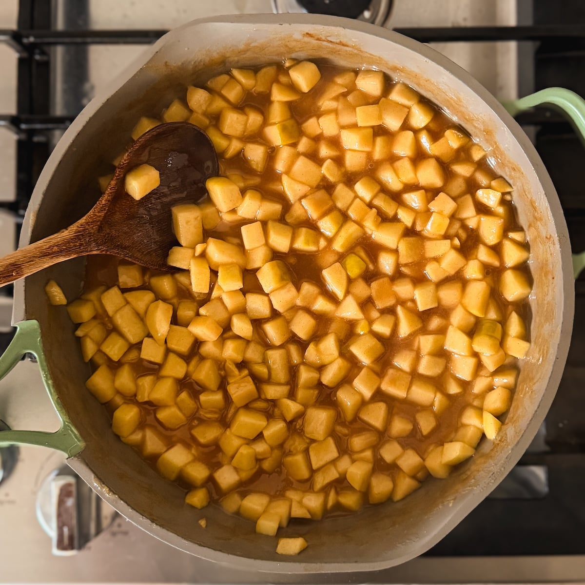 Large pot on a stove filled with diced apples cooking in a brown cinnamon sauce. A wooden spoon rests inside, stirring the mixture while the apples soften and caramelize.