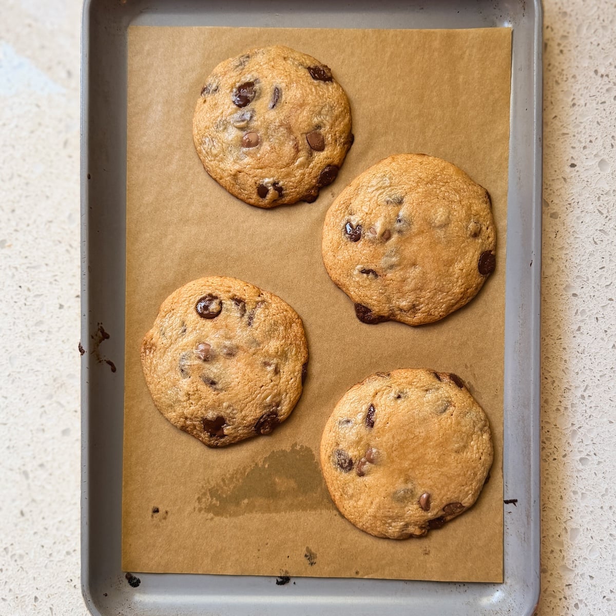 Four chocolate chip cookies on brown parchment paper atop a metal baking sheet, resting on a light-colored countertop.