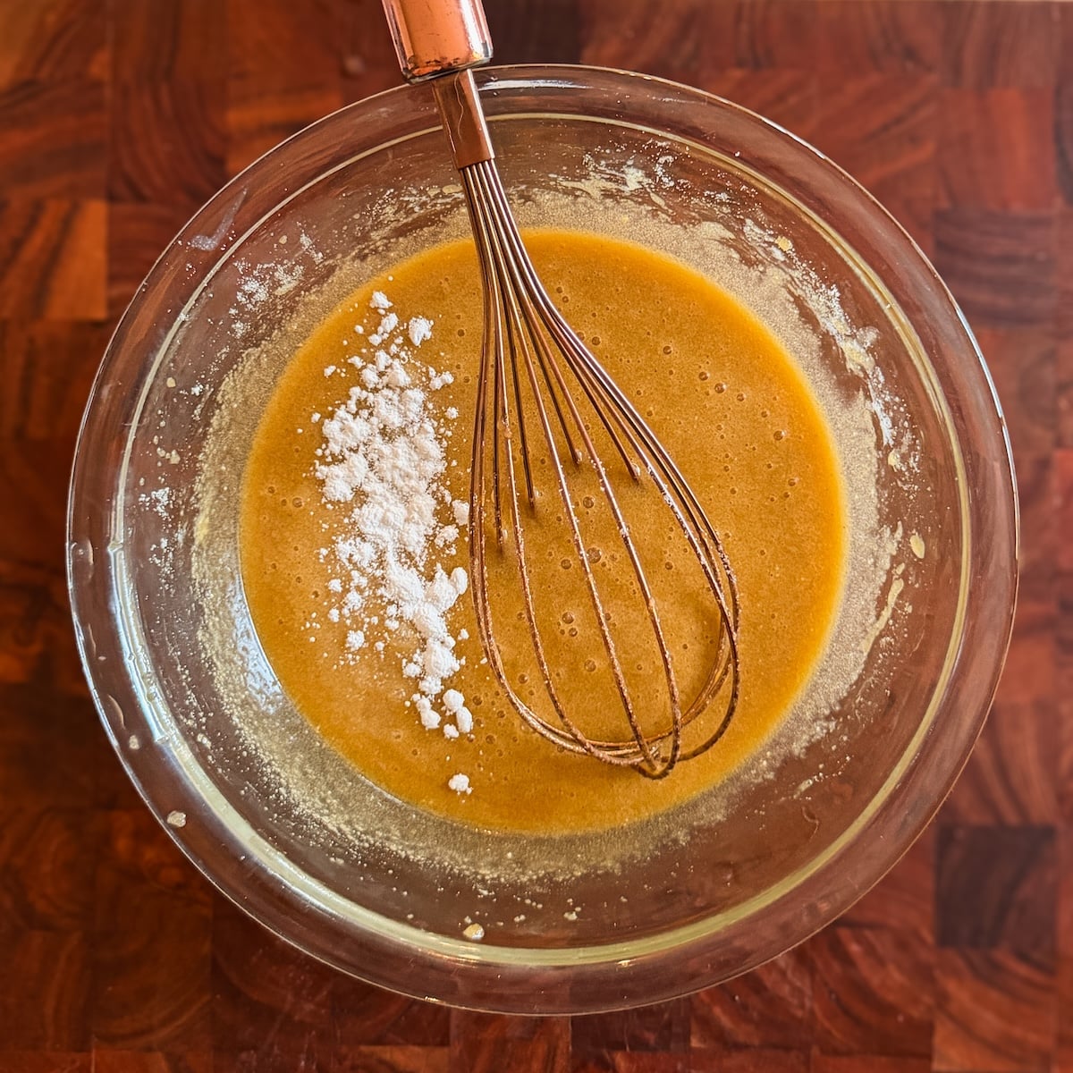 A glass bowl of light brown batter with a whisk and some baking powder and salt on top, sits on a wooden surface.