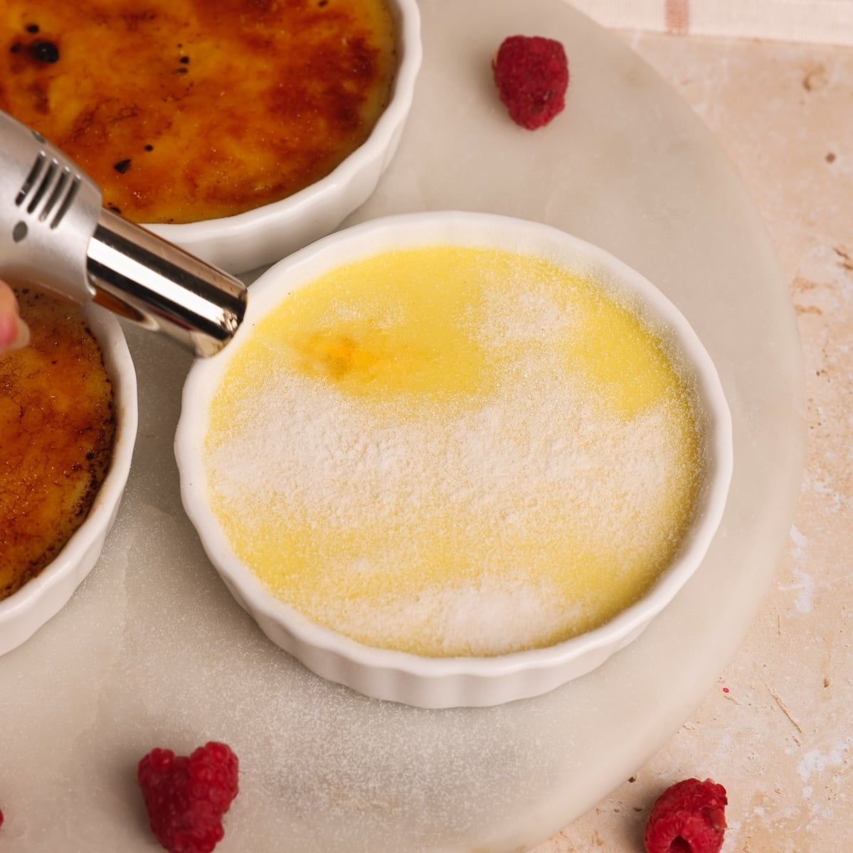 A hand holds a torch over a ramekin of crème brûlée, caramelizing sugar. Another finished crème brûlée and some raspberries are nearby on a marble surface.