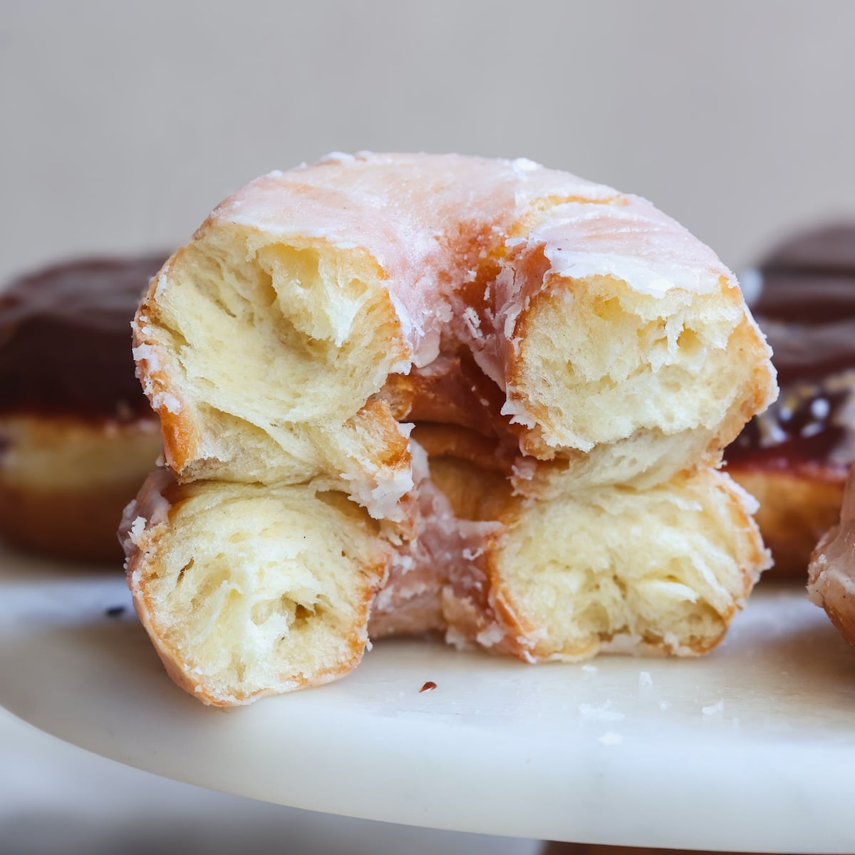 A close-up of a glazed donut torn in half, revealing its airy, flaky interior. The donut rests on a white surface with other donuts blurred in the background.