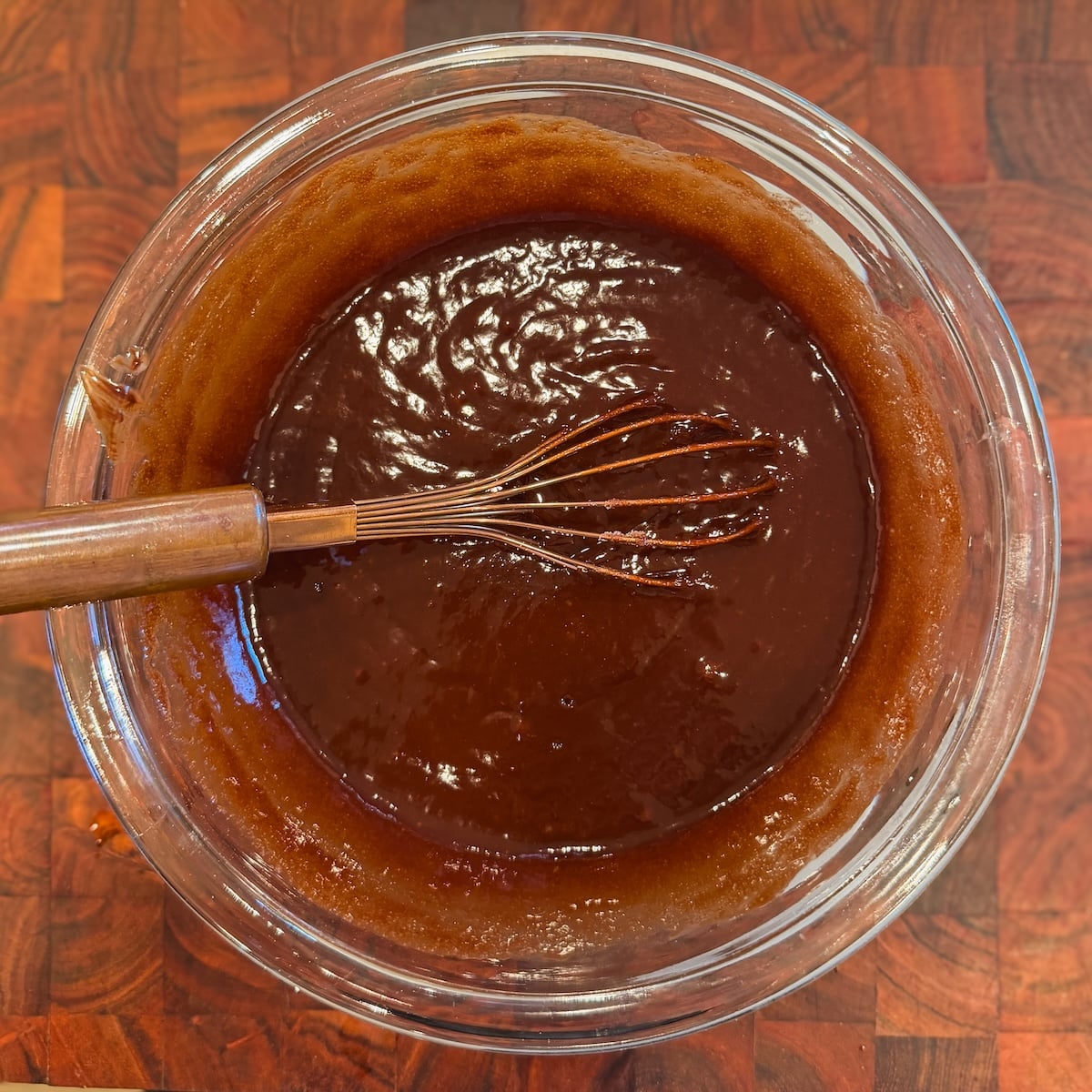 A glass bowl filled with melted chocolate brownie mixture and a whisk resting inside, placed on a wooden surface.