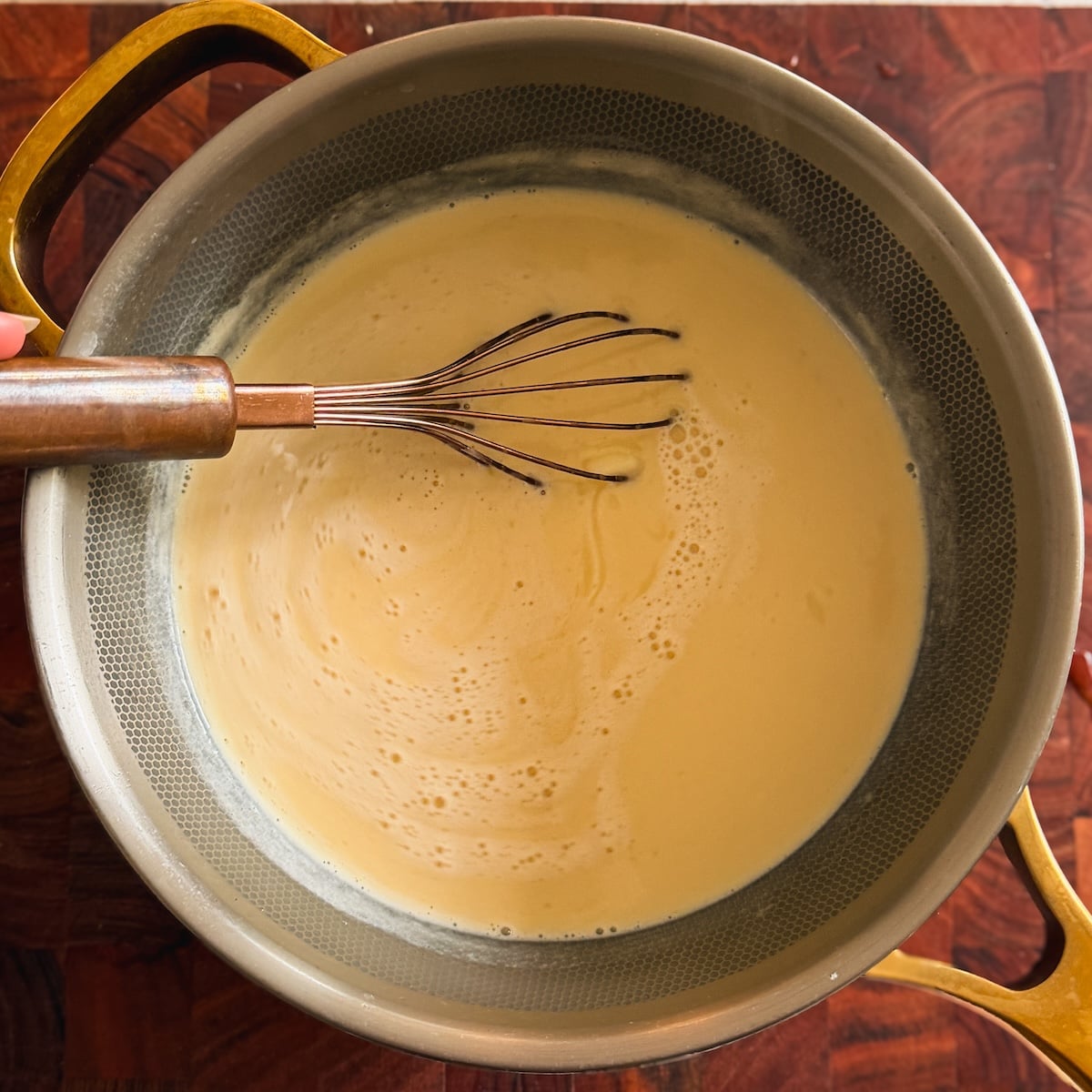 A metal whisk stirs a creamy custard in a saucepan with gold handles on a wooden surface.