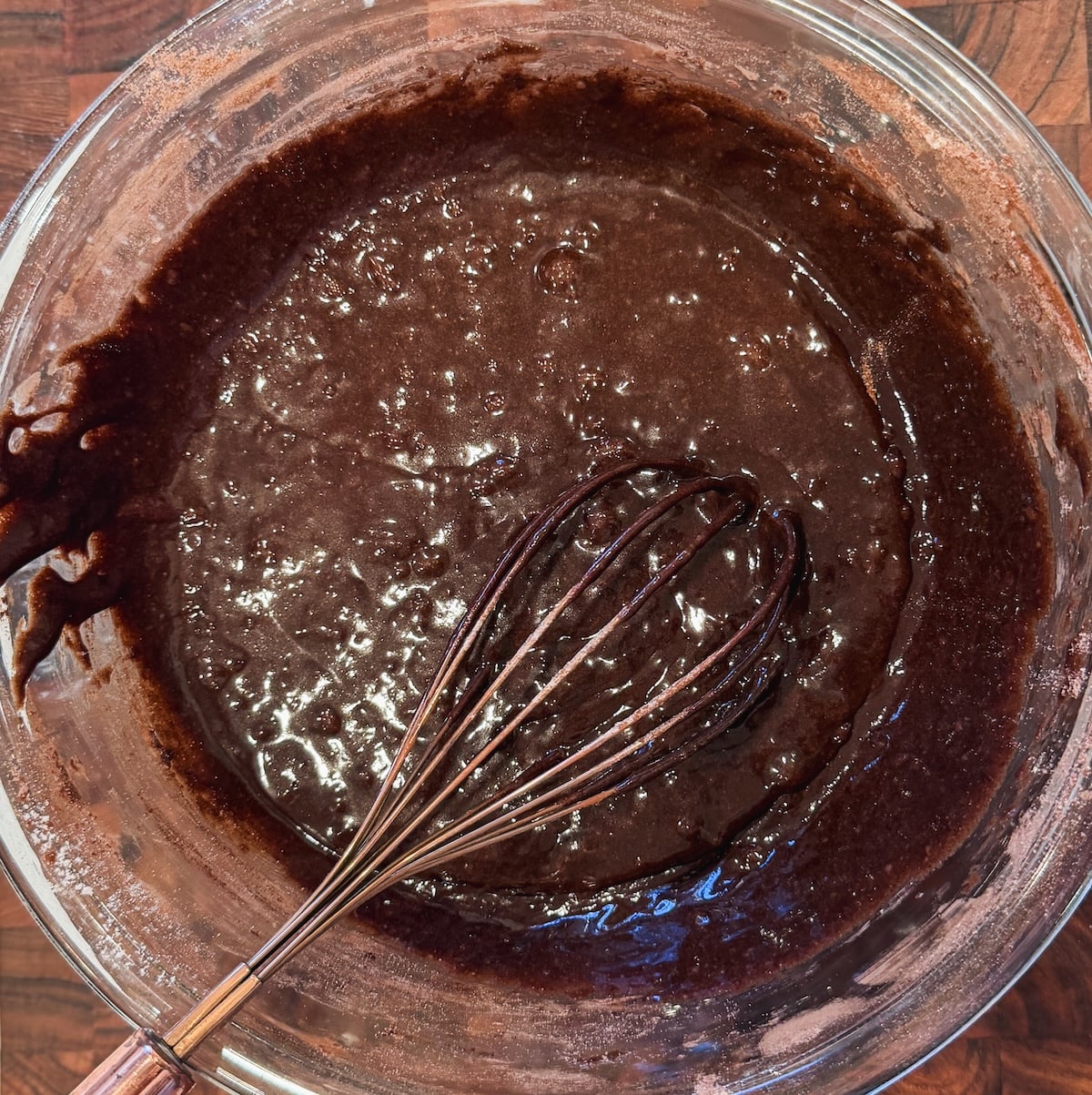 A glass bowl filled with brownie batter sits on a wooden surface, with a metal whisk partially submerged in the mixture.