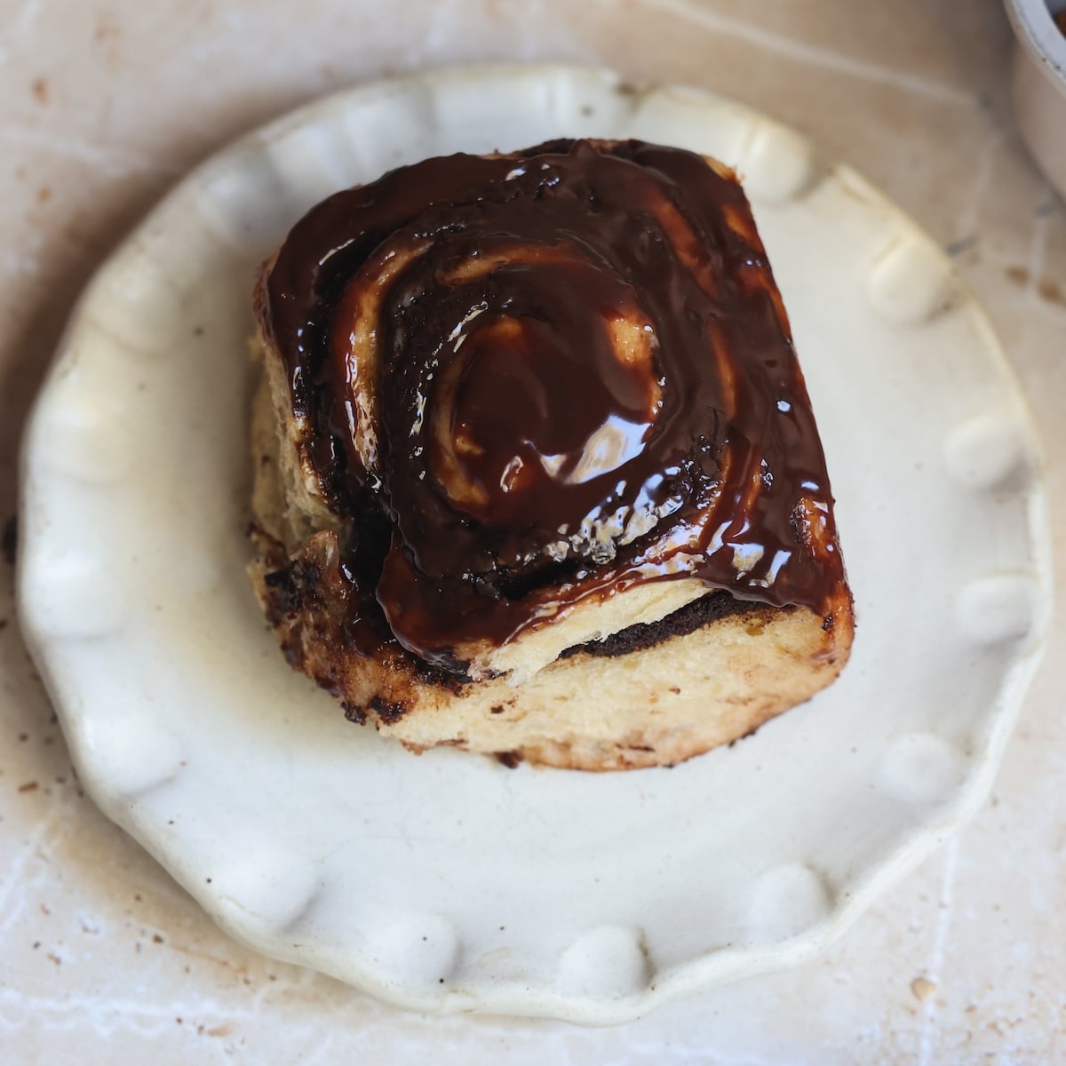 A single brownie cinnamon roll topped with glossy chocolate glaze sits on a white, scalloped-edge plate on a light-colored surface.