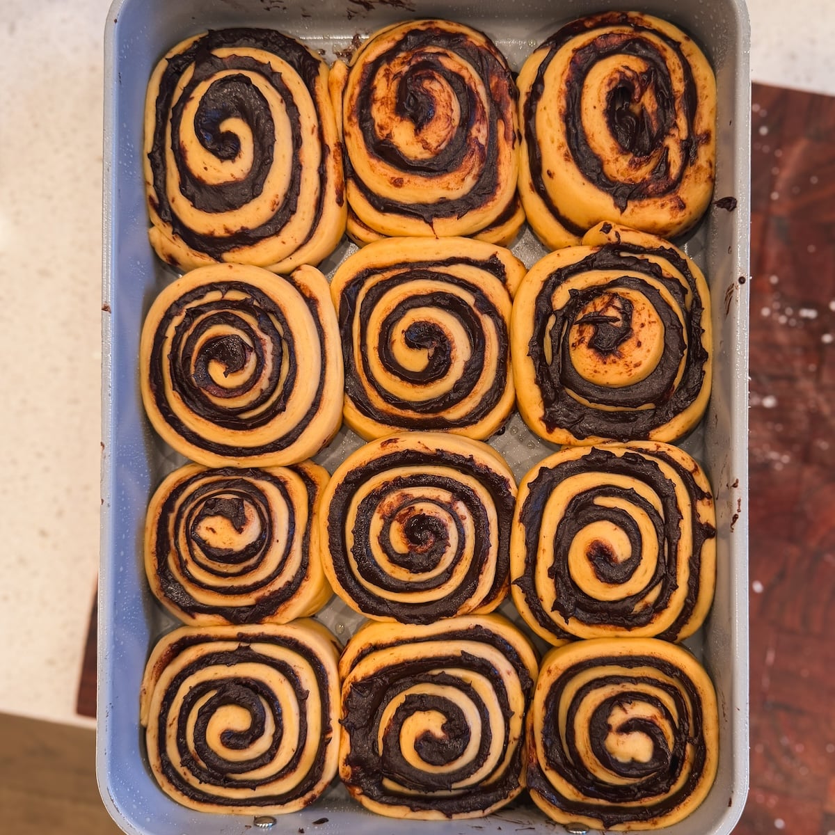 A baking tray filled with twelve unbaked chocolate brownie swirl rolls arranged in neat rows, showing a clear spiral pattern of chocolate filling in golden dough. Rolls look puffy