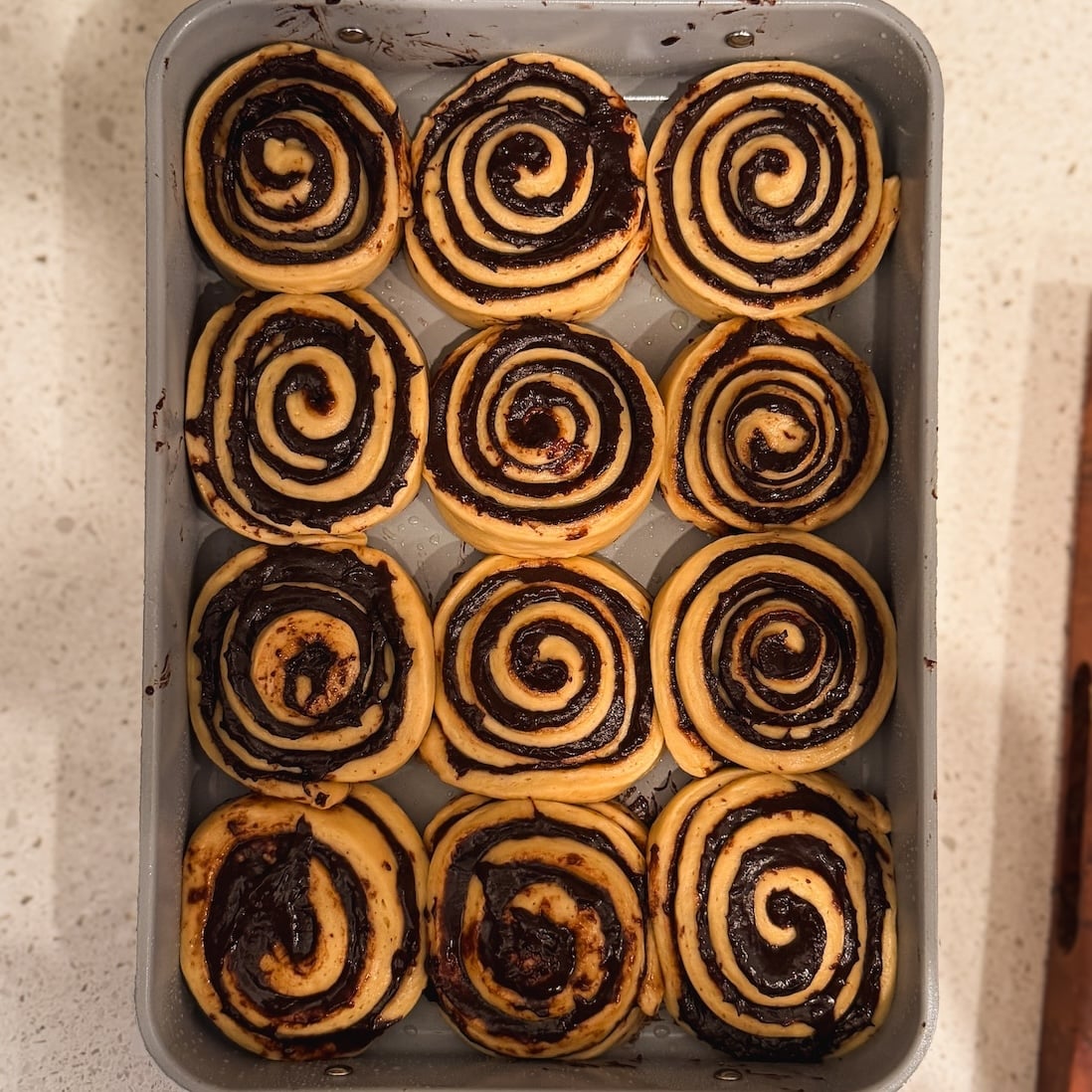 A baking tray with twelve unbaked chocolate swirl rolls arranged in three rows, sitting on a light-colored countertop.
