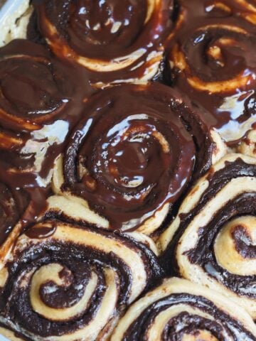 Close-up of freshly baked chocolate brownie swirl rolls in a baking tray, with some rolls generously topped with glossy chocolate glaze, while others remain unglazed.
