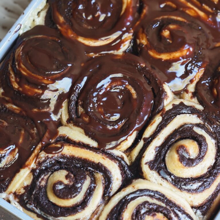 Close-up of freshly baked chocolate brownie swirl rolls in a baking tray, with some rolls generously topped with glossy chocolate glaze, while others remain unglazed.